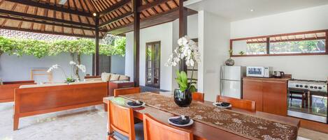 A dining area with a wooden table, chairs, and a ceiling fan. There is a kitchen area with a refrigerator and stove.