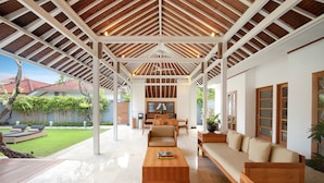 A covered outdoor living area with a wooden ceiling, a sofa, a coffee table, and a view of a garden and house in the background. - Uma Sapna Seminyak (Seminyak)