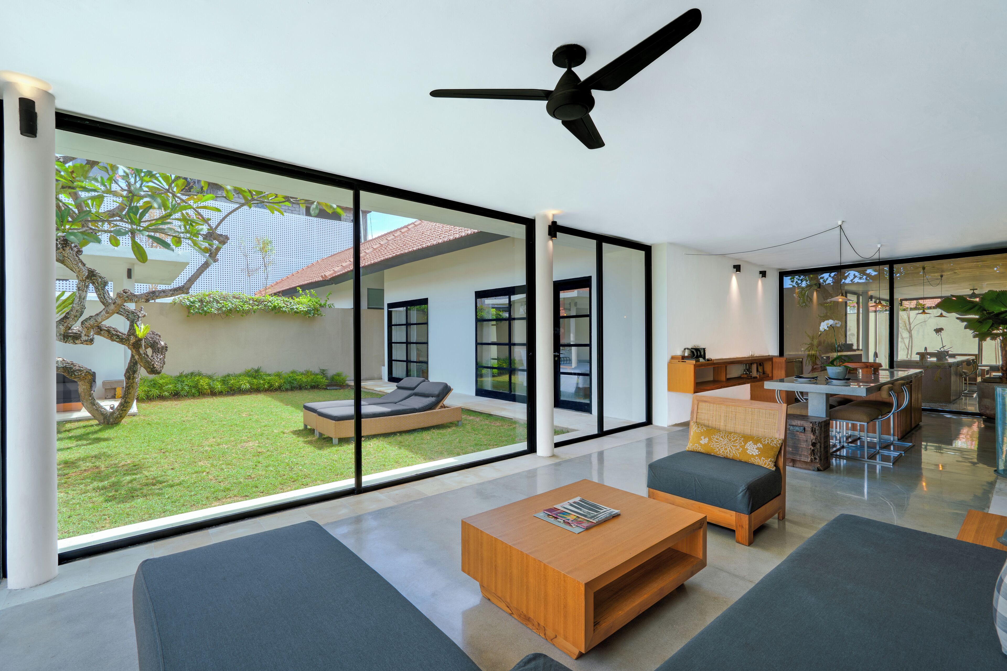 Un salon moderne doté de grandes portes vitrées donnant sur un jardin, d’un ventilateur de plafond et d’un coin repas avec une cuisine visible à travers le verre.