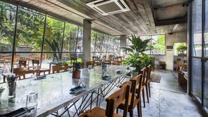 A spacious indoor dining area with a long table set for multiple guests, surrounded by large windows offering views of greenery. - Uma Sapna Seminyak (Seminyak)