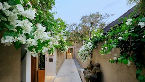 A narrow pathway flanked by walls adorned with white flowers and greenery, leading to a traditional entrance. - Uma Sapna Seminyak (Seminyak)