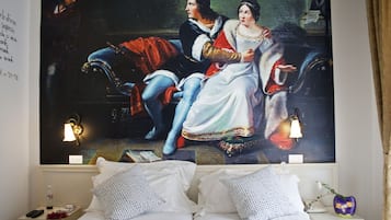 Ein Hotelzimmer mit einem großen Gemälde im klassischen Stil an der Wand, einem ordentlich bezogenen Bett mit weißer Bettwäsche und einem Nachttisch mit Lampe und Blumenvase.