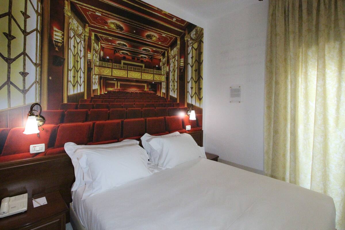 Una habitación de hotel con una cama grande, una mesita de noche con un teléfono y un mural en la pared de un teatro.
