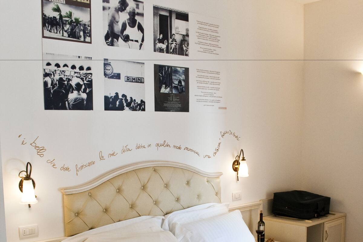 Una cama bien hecha con ropa blanca, una mesita de noche con una lámpara, y una pared decorada con fotografías y texto en blanco y negro.