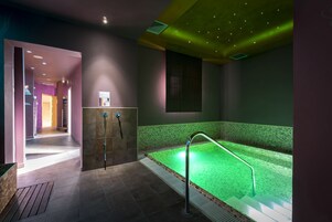 Un'area spa con vasca idromassaggio illuminata di verde, zona doccia e corridoio che conduce ad altre stanze.