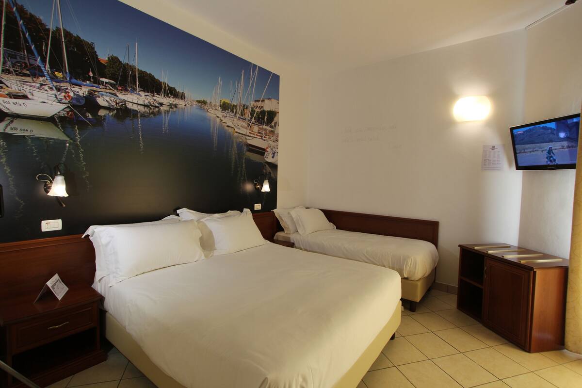 Habitación de hotel con dos camas, televisión, luz empotrada en la pared y un mural grande de un puerto deportivo.