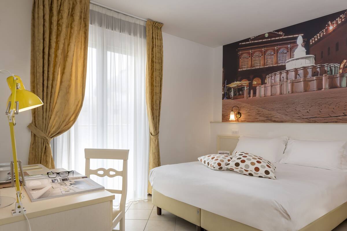 Habitación de hotel con cama, un escritorio con lámpara, una silla y un cartel grande de un edificio.