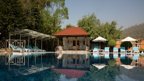 Una piscina con sillas de descanso y sombrillas, rodeada de árboles y un pabellón.