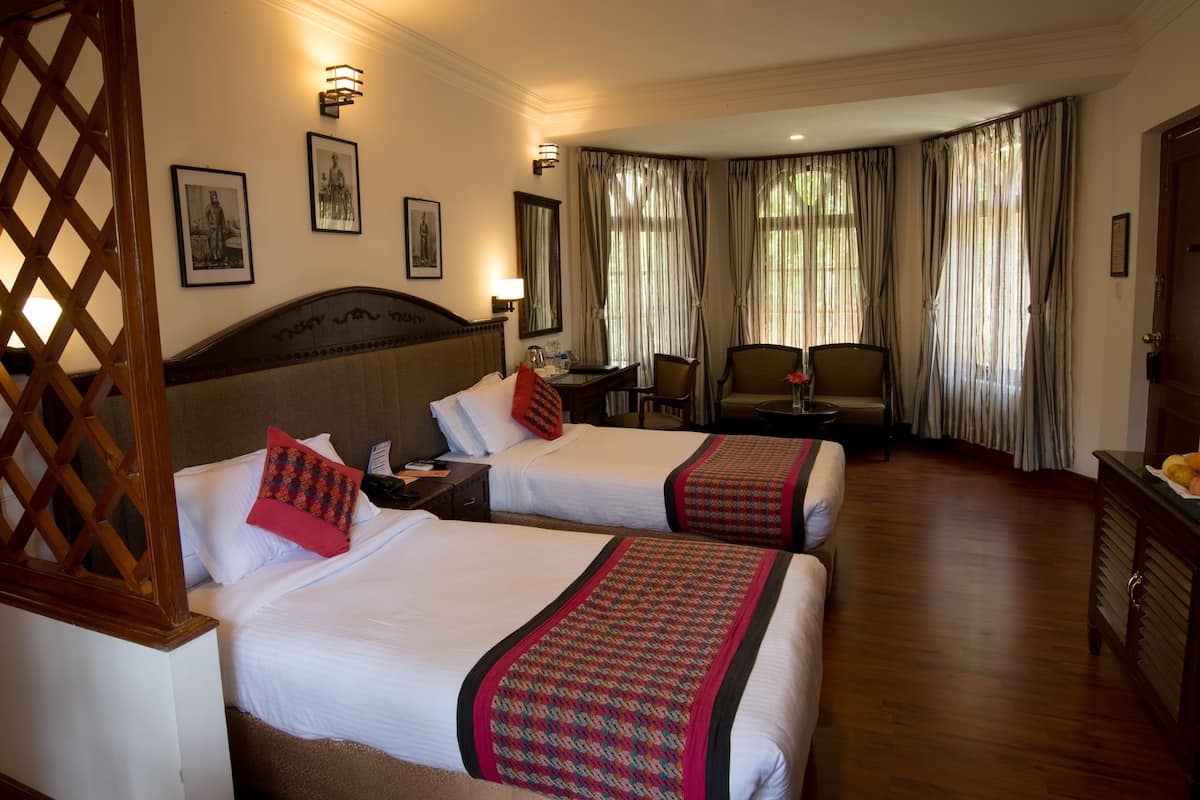 Quarto de hotel com duas camas, uma escrivaninha, uma cadeira e um sofá. Quadros emoldurados adornam a parede e há uma janela com cortinas.