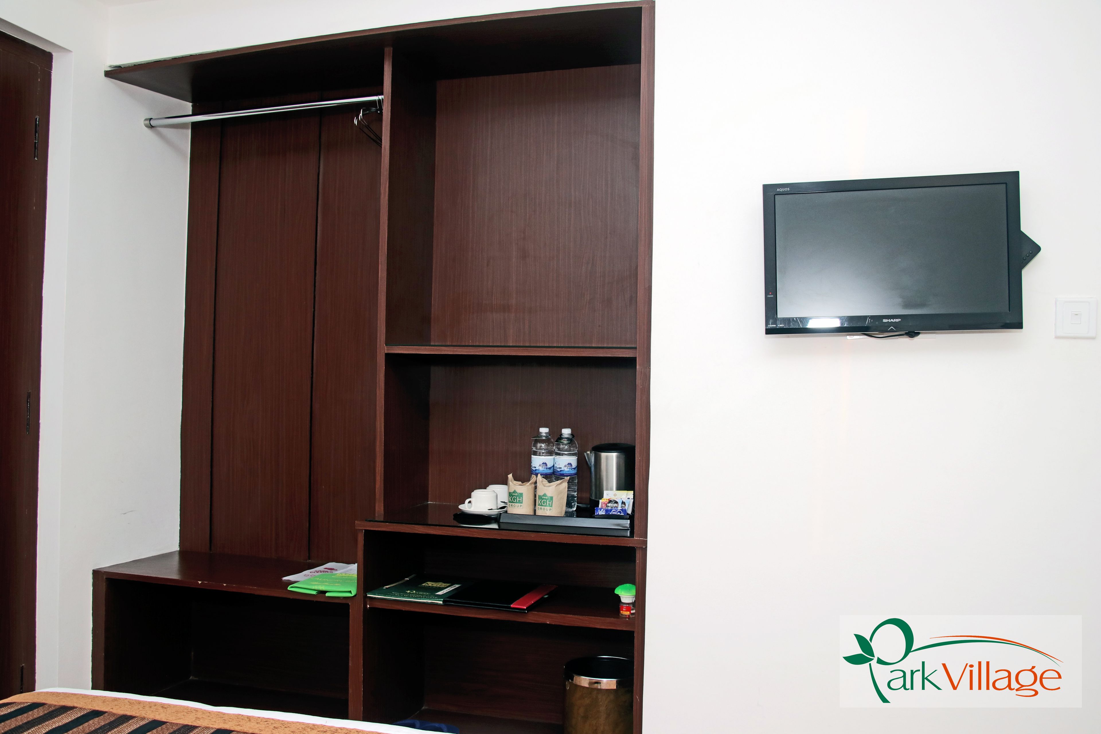 A room with a wardrobe, a television mounted on the wall, and a shelf with various items including a kettle, cups, and a bag.