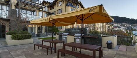 An outdoor patio with wooden tables and benches, a grill, and a yellow umbrella.