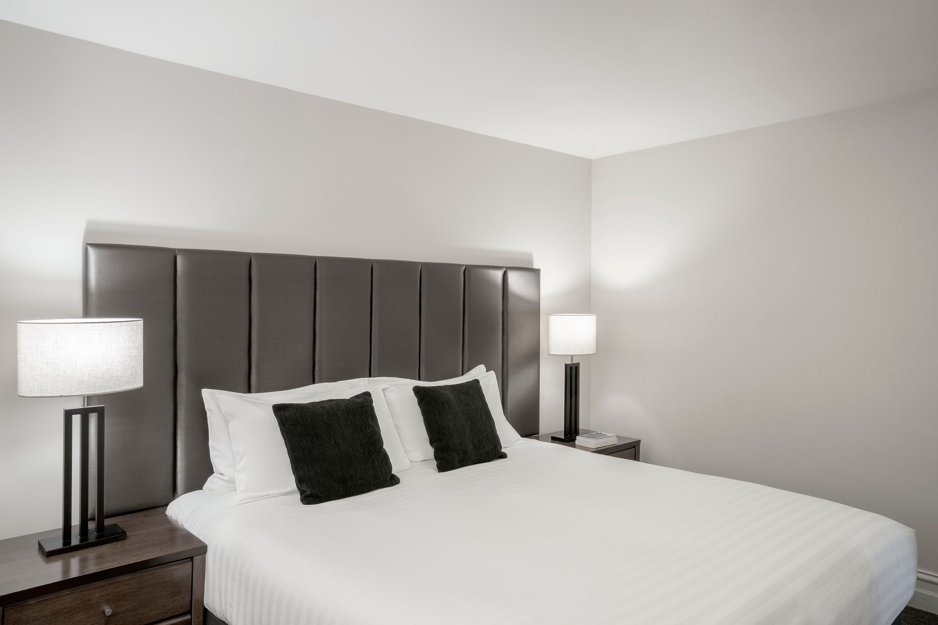 A neatly made bed with white linens and two black pillows, flanked by two bedside tables with lamps, in a modern hotel room.