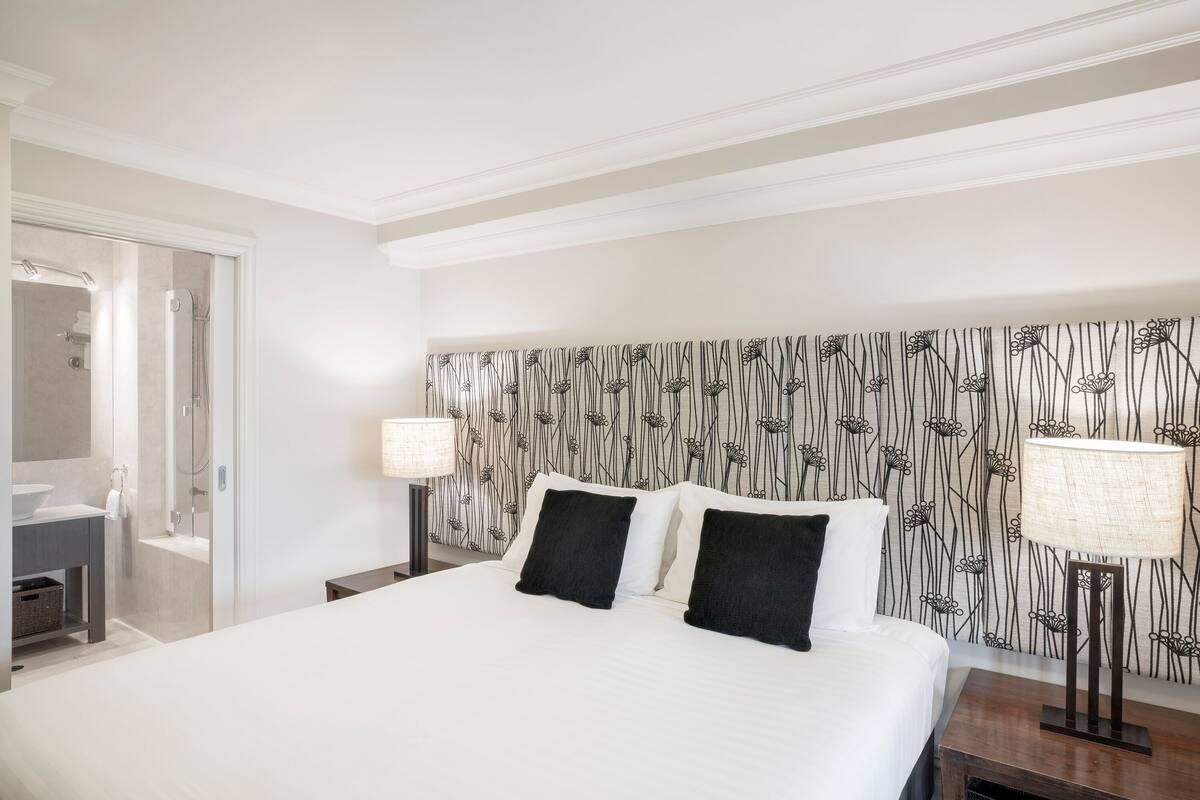 Hotellihuone, jossa on suuri sänky, yöpöytä lampun kanssa, kylpyhuone, jossa on suihku ja pesuallas, sekä kuviollinen tapetti seinällä.