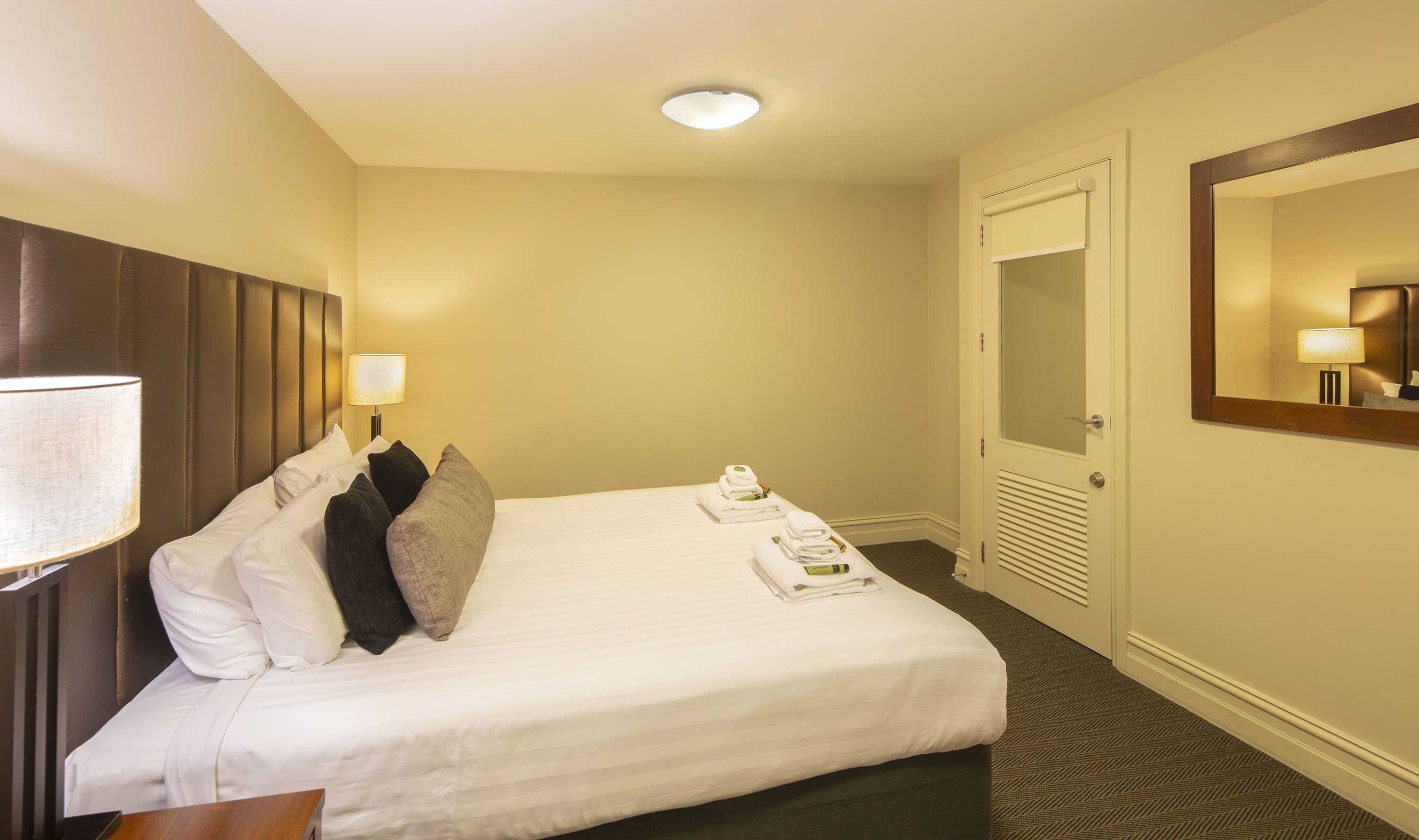 A hotel room with a neatly made bed, a bedside lamp, a mirror, and a door with a window.