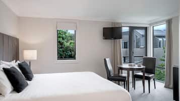 A hotel room with a large bed, a small table with chairs, a TV, and a balcony with a view of greenery.