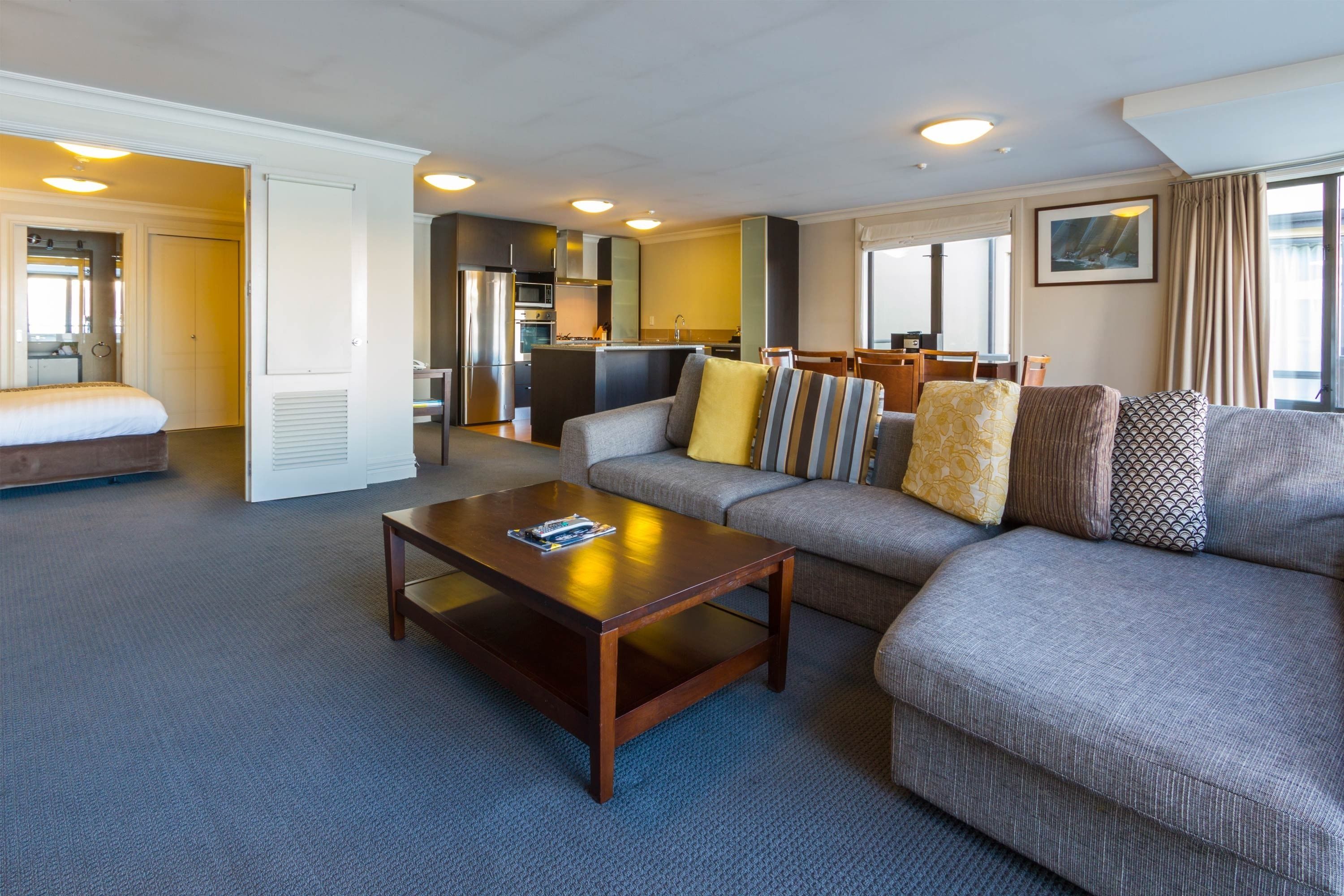 A modern hotel room with a living area, a sofa with patterned cushions, a wooden coffee table, and a kitchen in the background.