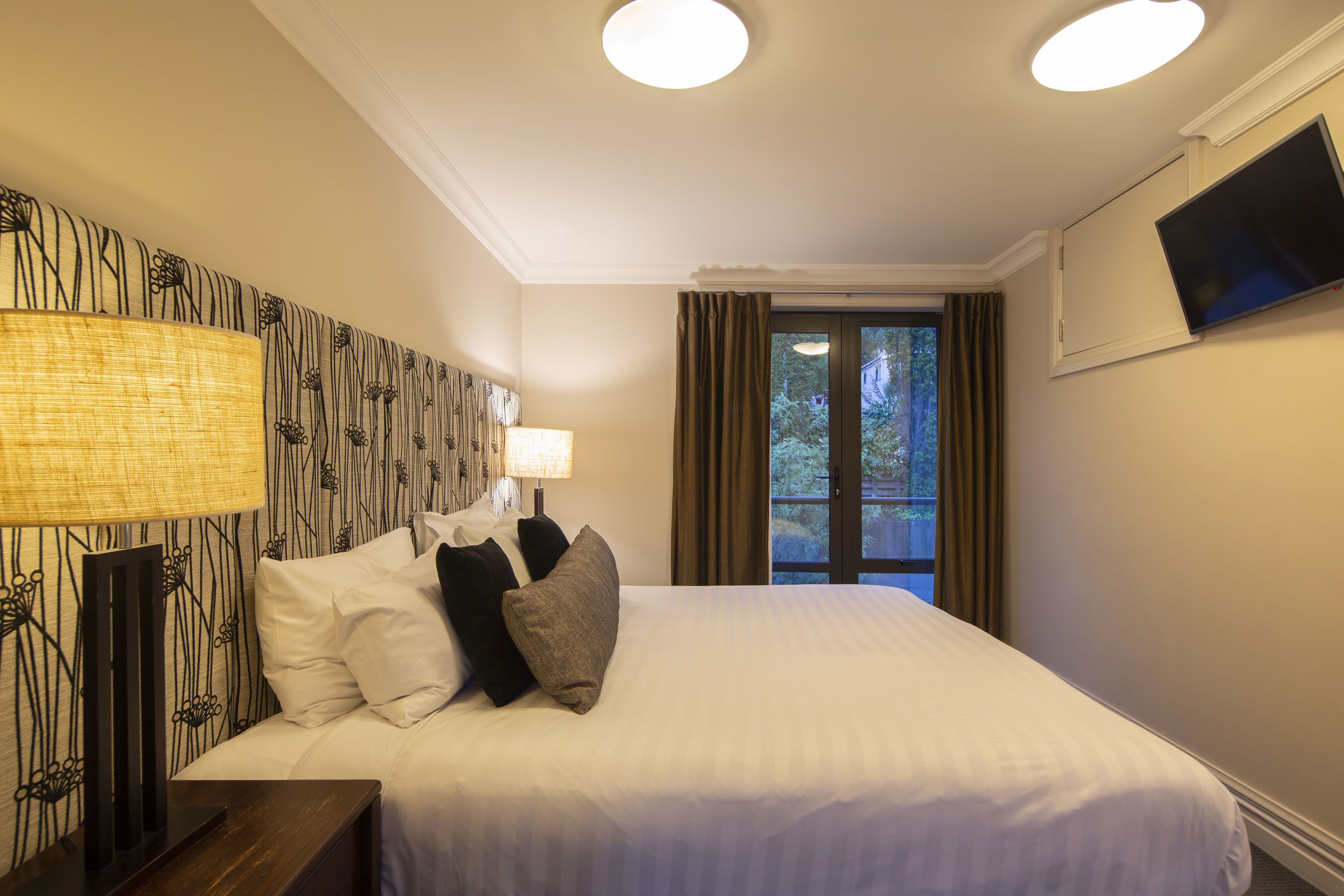 A hotel room with a large bed, a nightstand, a lamp, a TV, and a window with curtains.