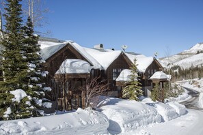 Una cabaña cubierta de nieve con varias cabañas de madera, cielo despejado y una montaña nevada al fondo.