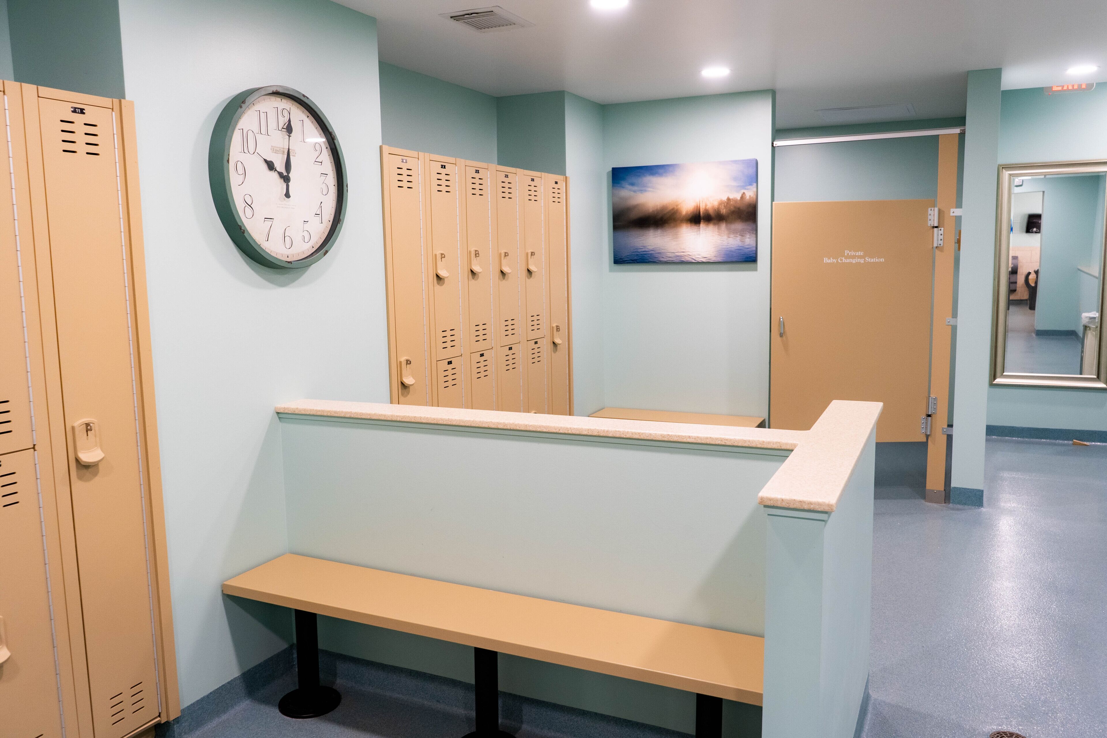A locker room with beige lockers, a clock, a bench, and a door labeled "Private Baby Changing Station."