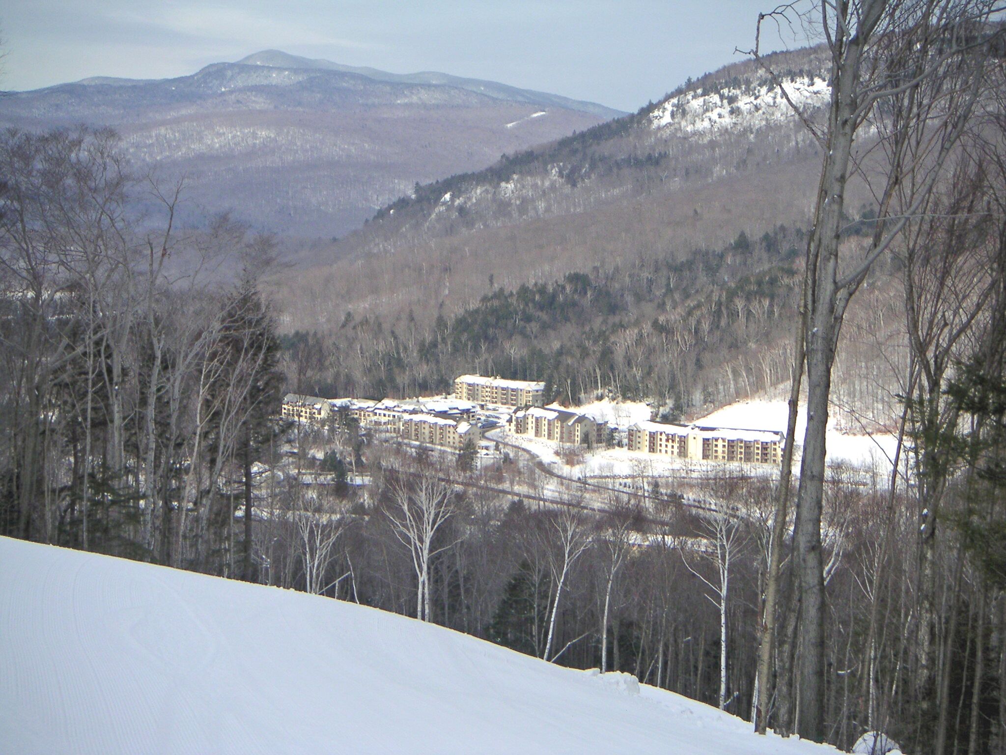 Un paysage enneigé avec un ensemble de bâtiments, probablement un hôtel, entouré d’arbres et avec des montagnes en arrière-plan.