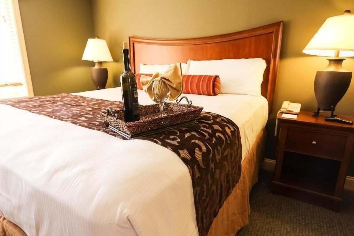Una habitación de hotel con una cama, una mesita de noche, una lámpara, una botella y un vaso.