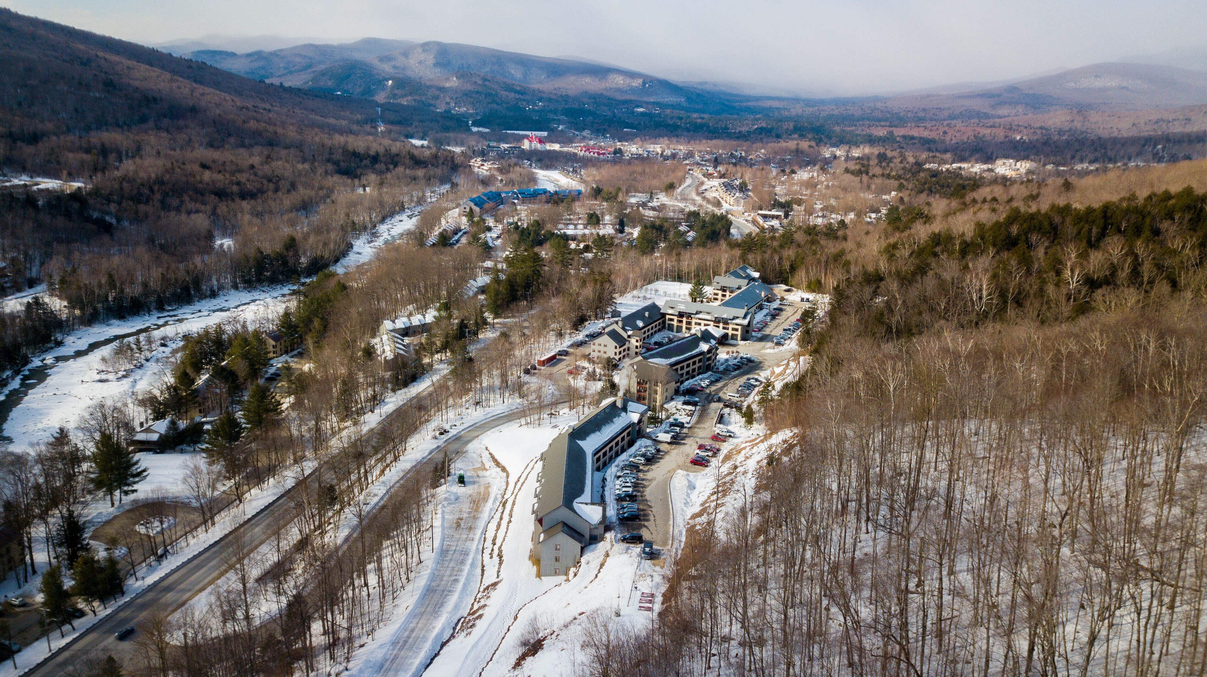 An aerial view of a resort with multiple buildings, a parking lot, and a snow-covered landscape with mountains in the background.