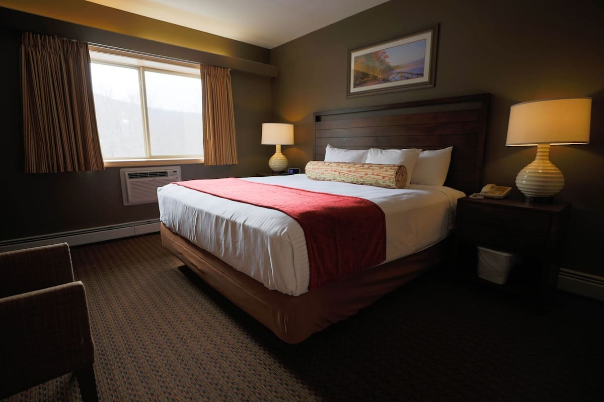 A hotel room with a large bed, two bedside lamps, a window with curtains, a painting on the wall, and a chair.