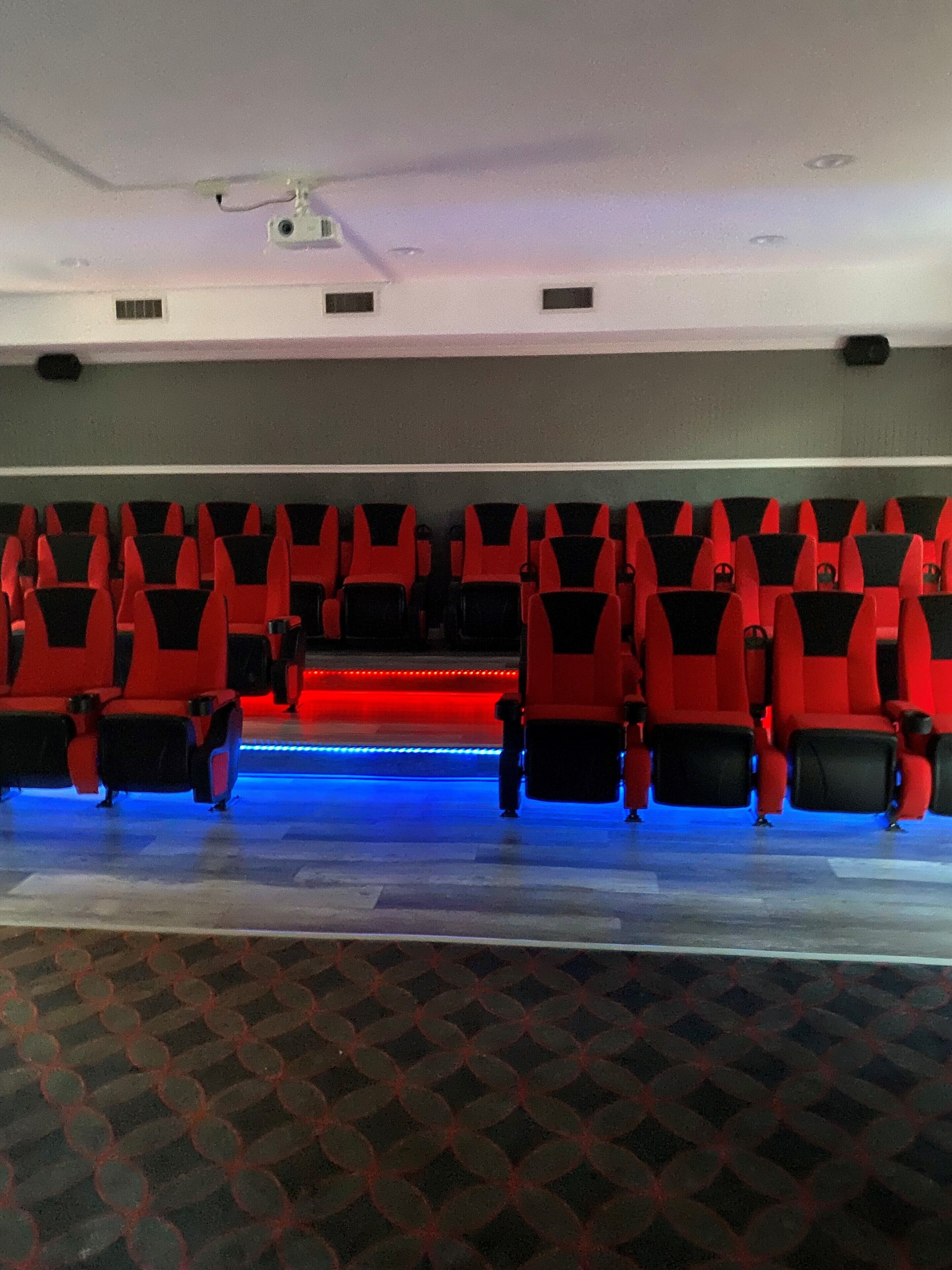 A theater with rows of red seats, a stage with blue lighting, and a ceiling projector.