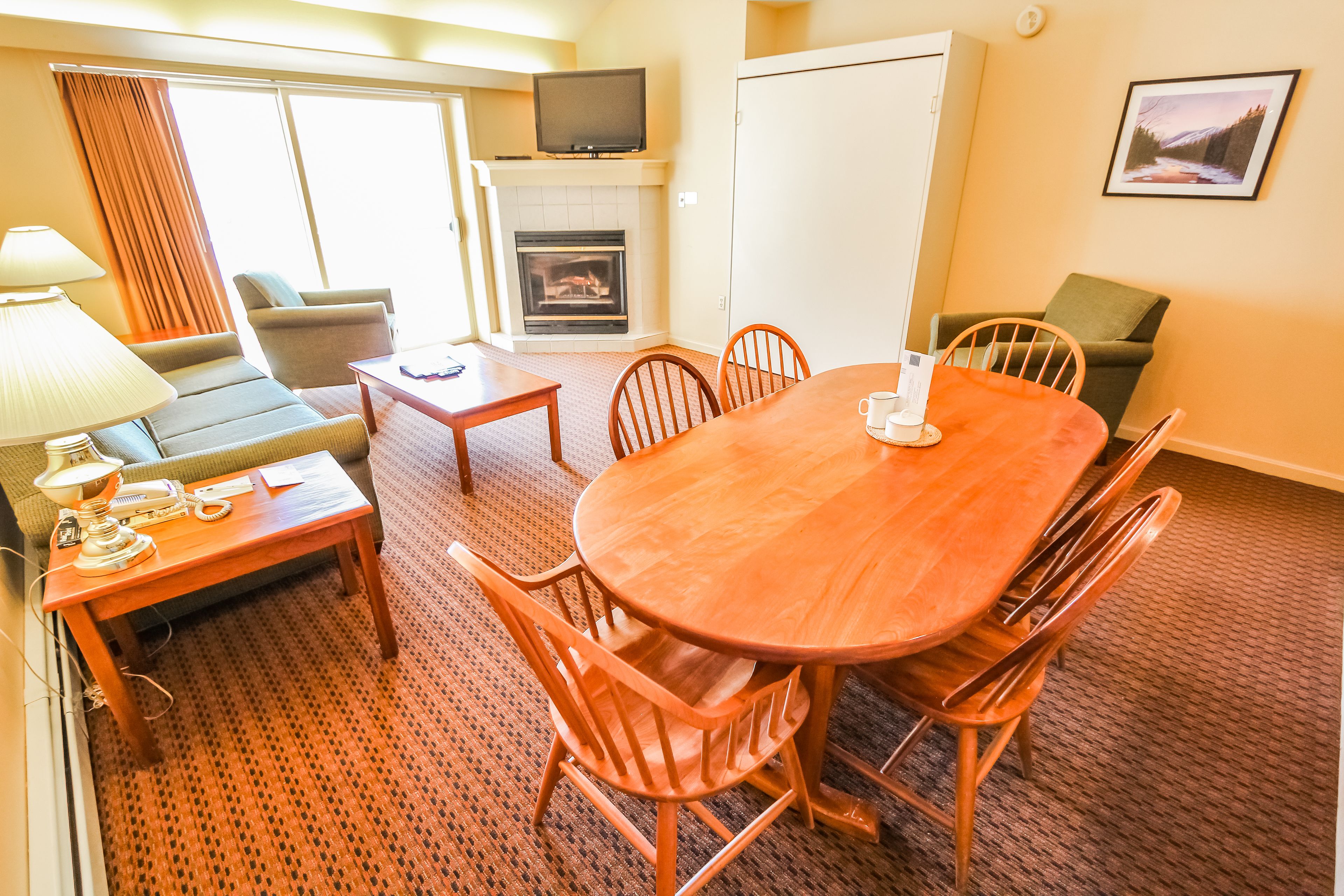 A hotel room with a dining table, chairs, a sofa, a TV, a fireplace, and a carpeted floor.