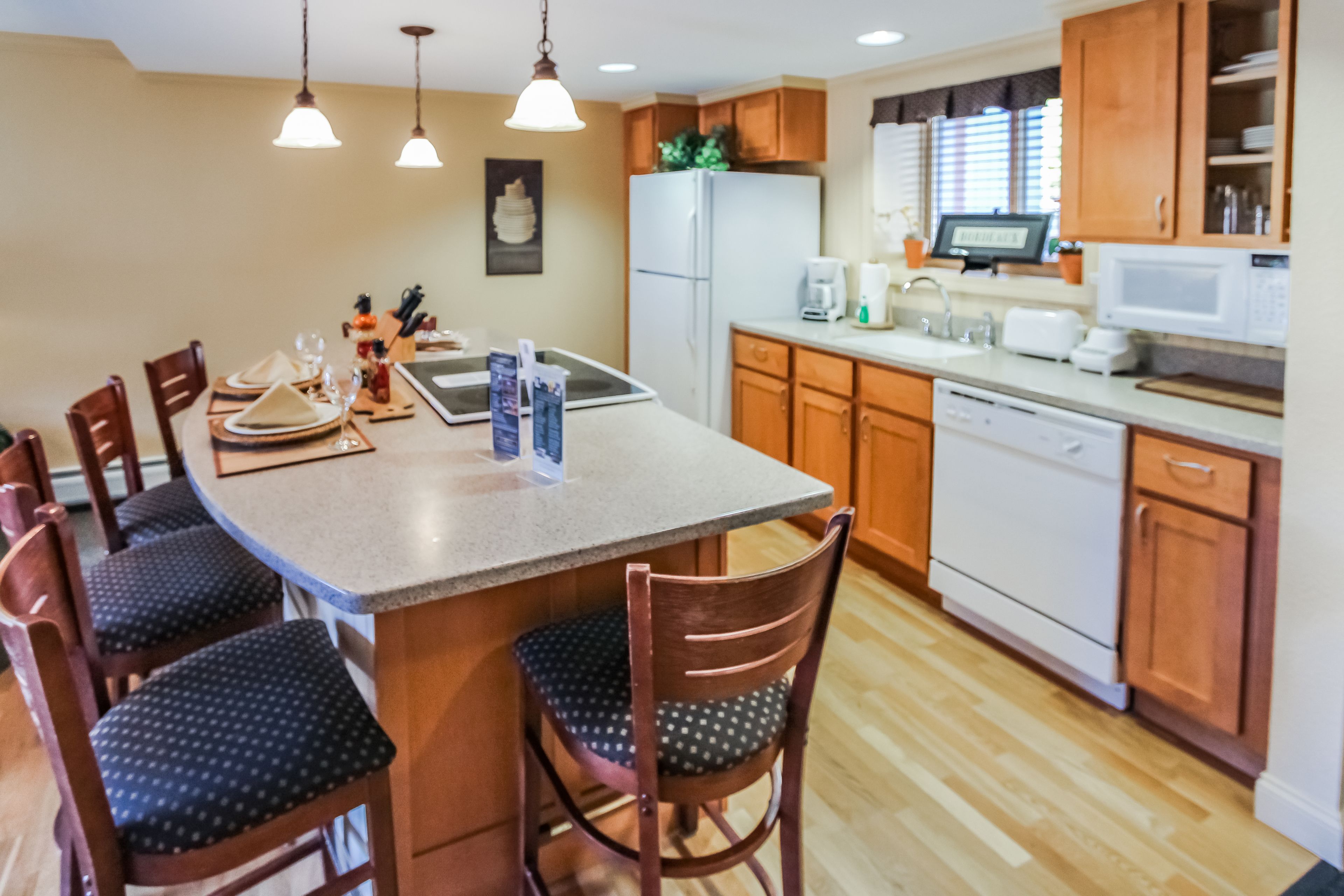 A kitchen with wooden cabinets, a white refrigerator, a microwave, and a dishwasher. There are two pendant lights hanging above the island.