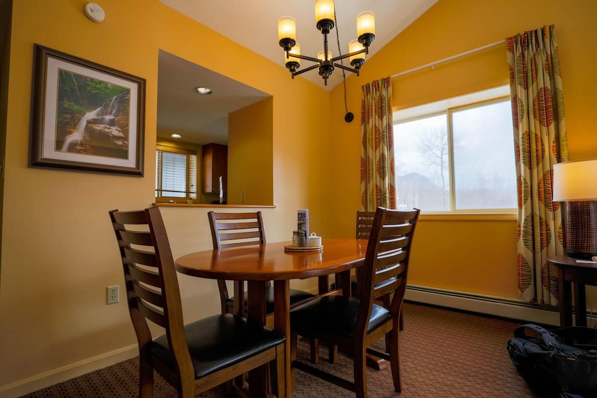A dining area with a round table, four chairs, a chandelier, a framed picture on the wall, and a window with curtains.