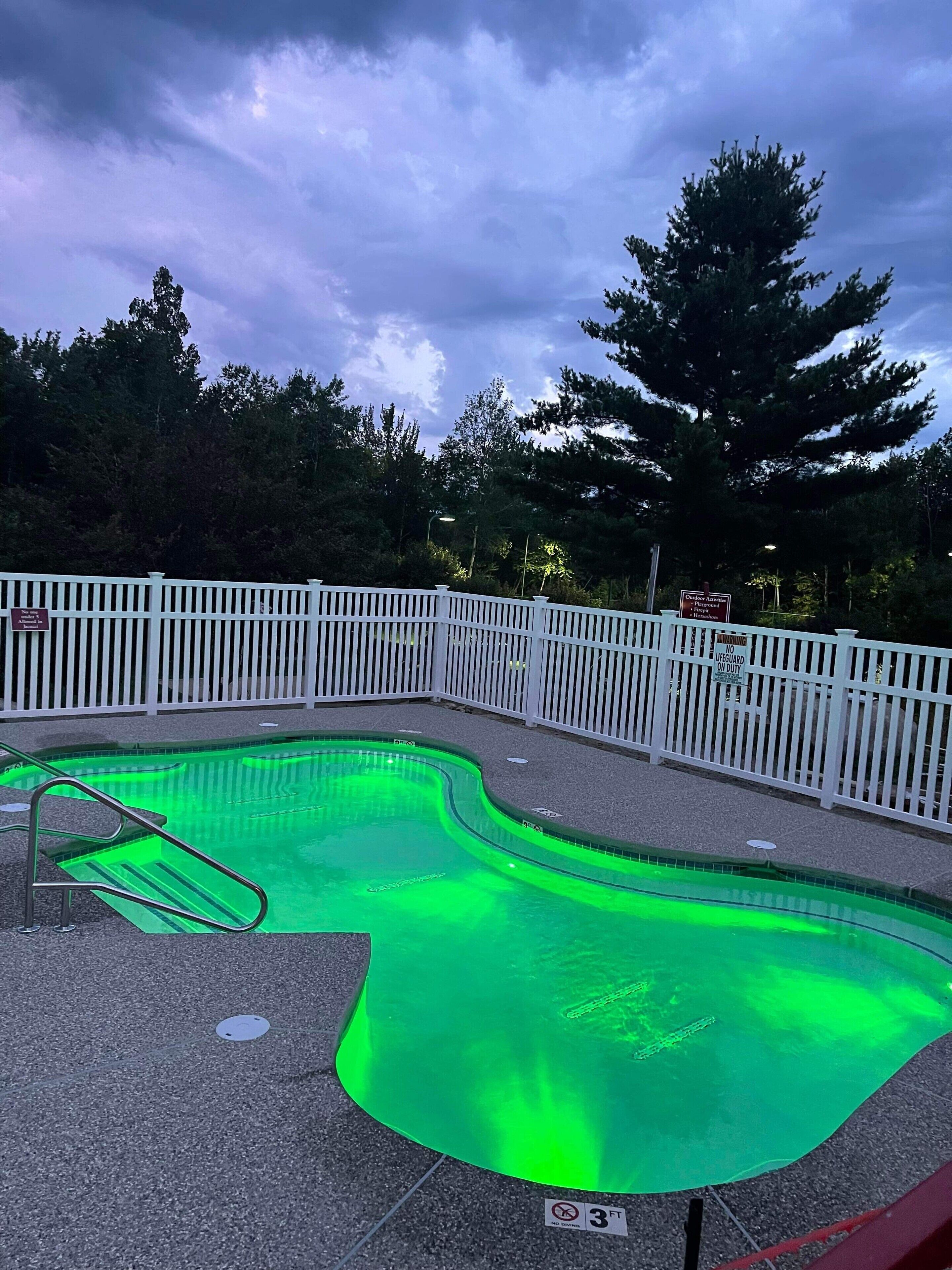 A swimming pool with a white fence, surrounded by trees and a cloudy sky.