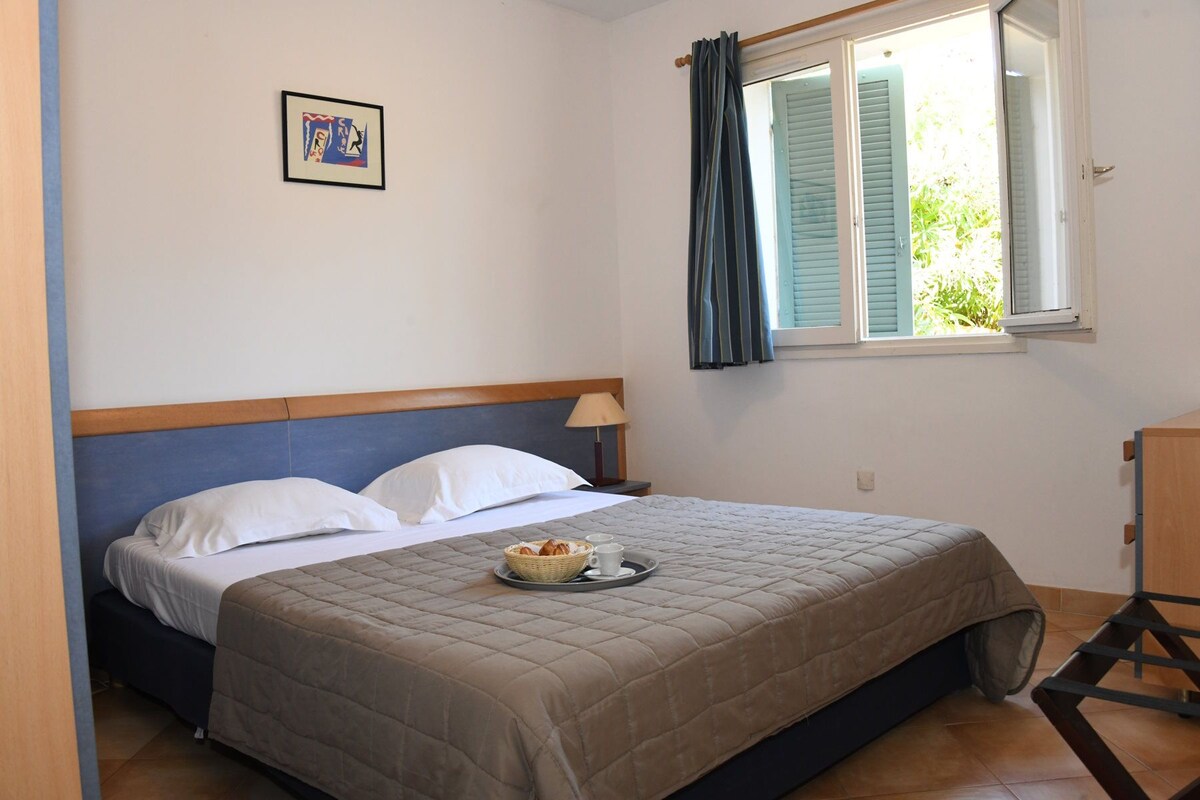 Un dormitorio con cama, una mesita de noche con lámpara, una ventana con cortinas y un cuadro en la pared.