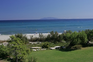 A beach with clear blue water, a distant mountain, and green vegetation.