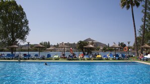 Piscina com espreguiçadeiras e guarda-sóis, rodeada por árvores e edifícios ao fundo.
