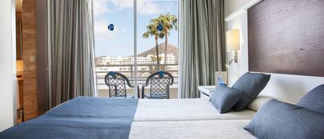 Habitación de hotel con una cama grande, una mesita de noche, una lámpara y vistas a palmeras y edificios.
