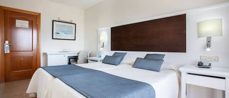 A hotel room with a large bed, a desk, a chair, a wooden door, a framed picture on the wall, and a nightstand with a lamp.