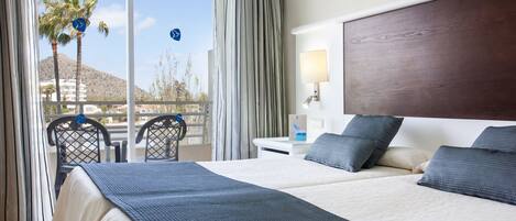A hotel room with two beds, a balcony with a view of palm trees and mountains, a desk with a lamp, and a chair.