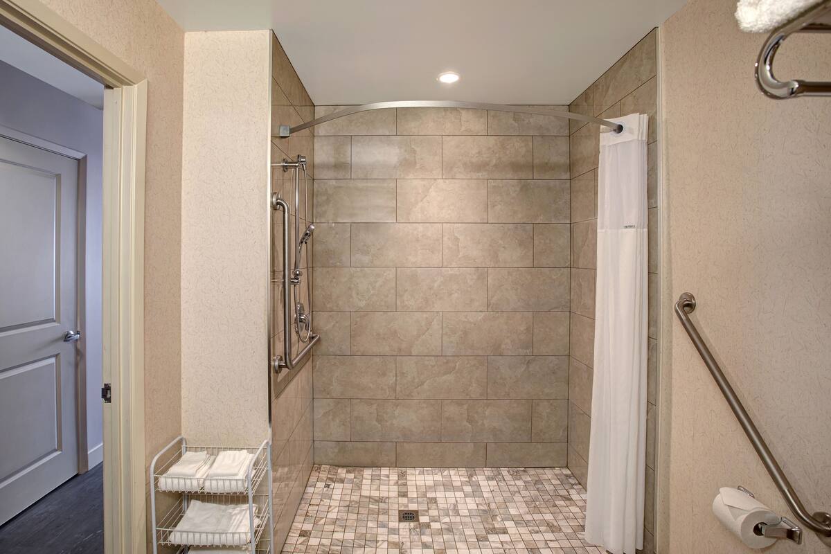 A bathroom with beige tiles, a shower with a white curtain, a grab bar, and a toilet paper holder.