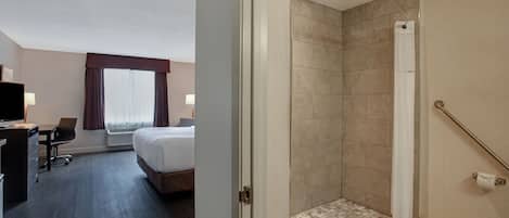 A hotel room with a bed, desk, and chair, a shower area with tiled walls, and a view of the outdoors through a window.