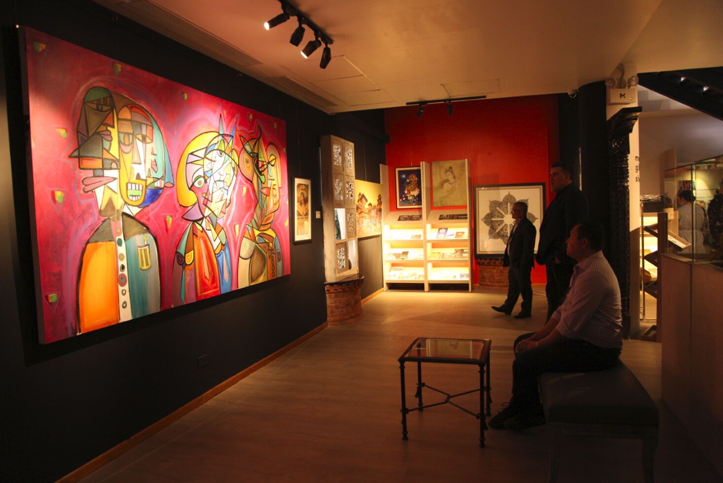 An art gallery with a large colorful painting on the wall, a glass display case, and a few people observing the artwork.