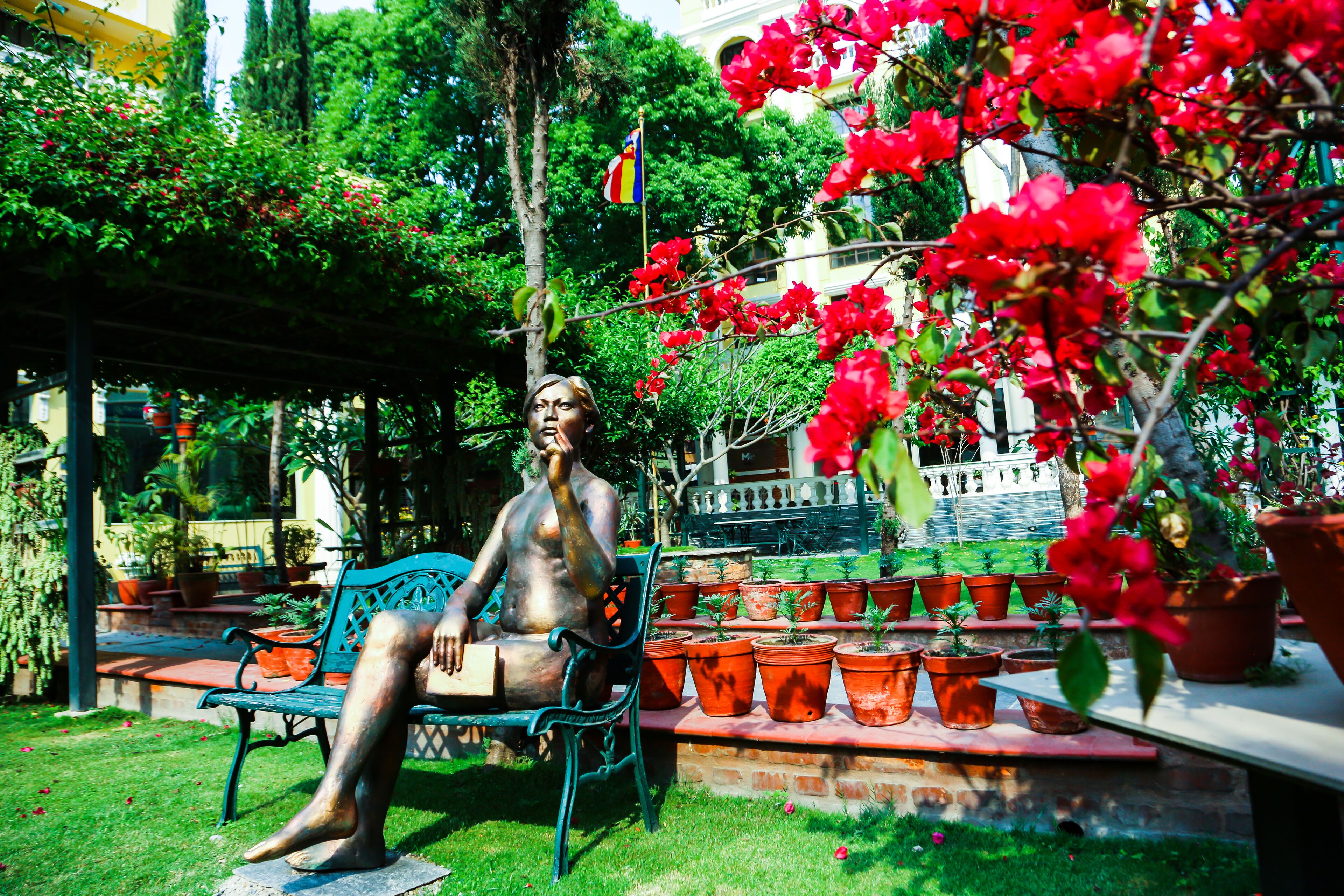 A statue of a seated figure on a bench in a garden with potted plants and a flag.