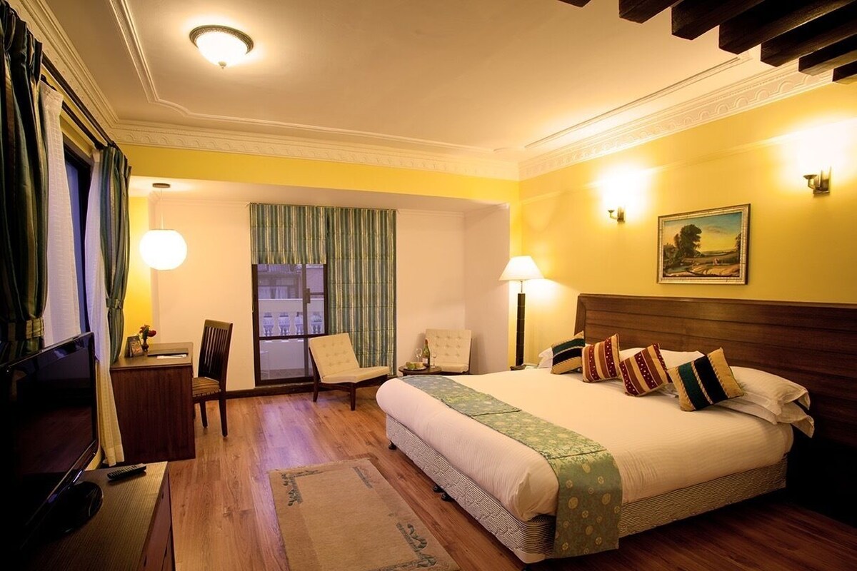 A hotel room with a large bed, two bedside lamps, a desk with a chair, a television, and a window with curtains.