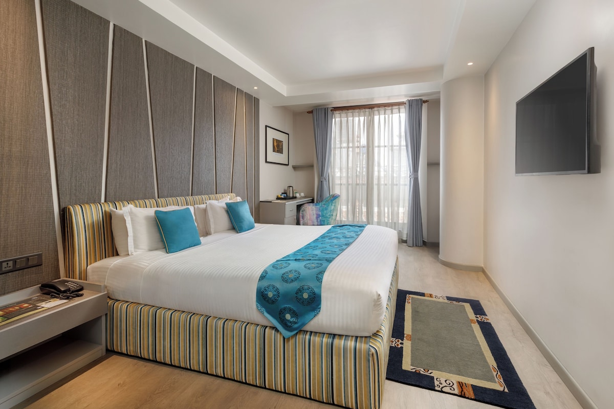 A hotel room with a large bed, a flat-screen TV, a bedside table with a phone, and a window with curtains.