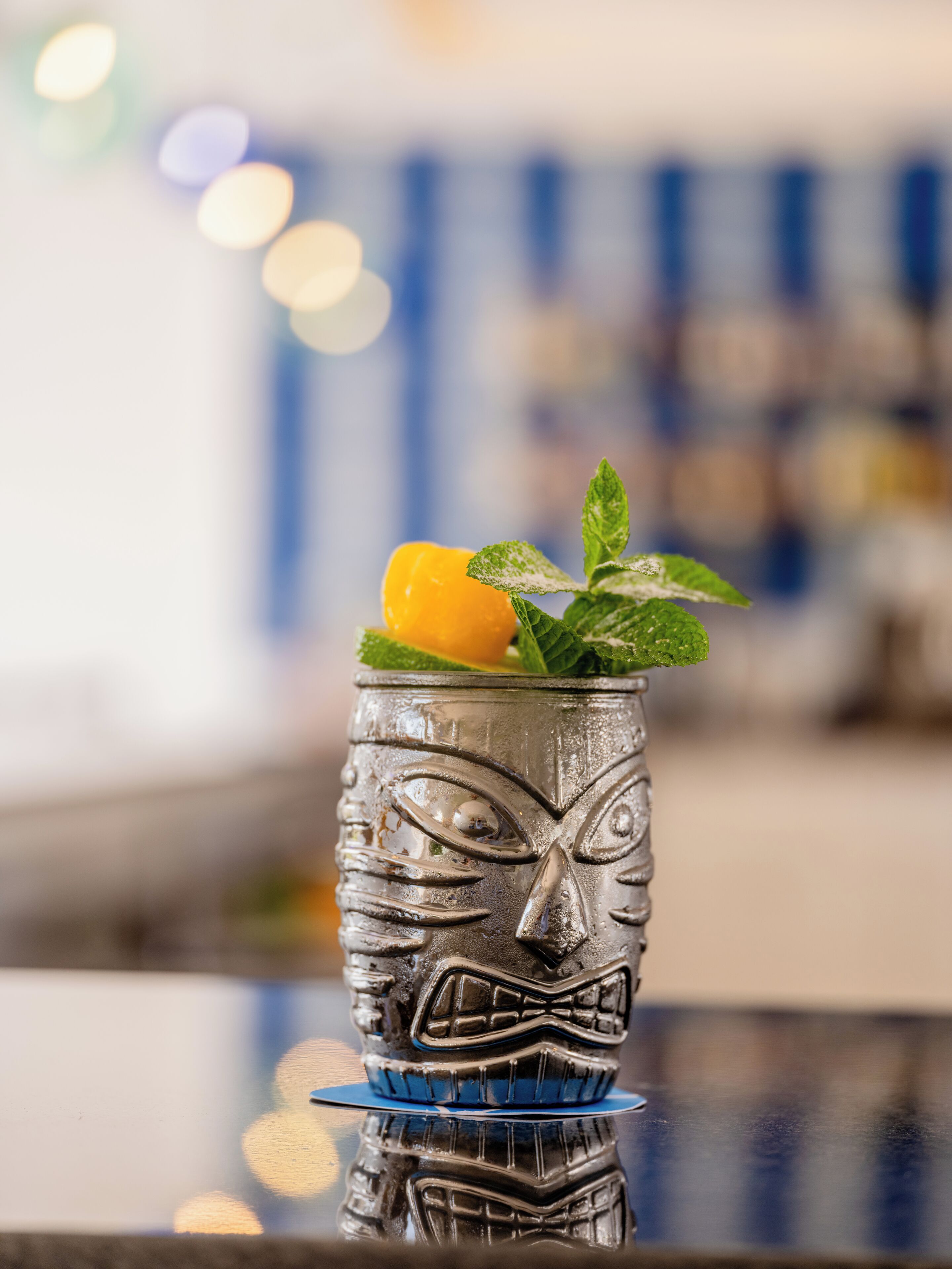 Ein Cocktailglas mit Gesichtsdekor, garniert mit einer Zitronenscheibe und Minzblättern, auf einer spiegelnden Oberfläche.