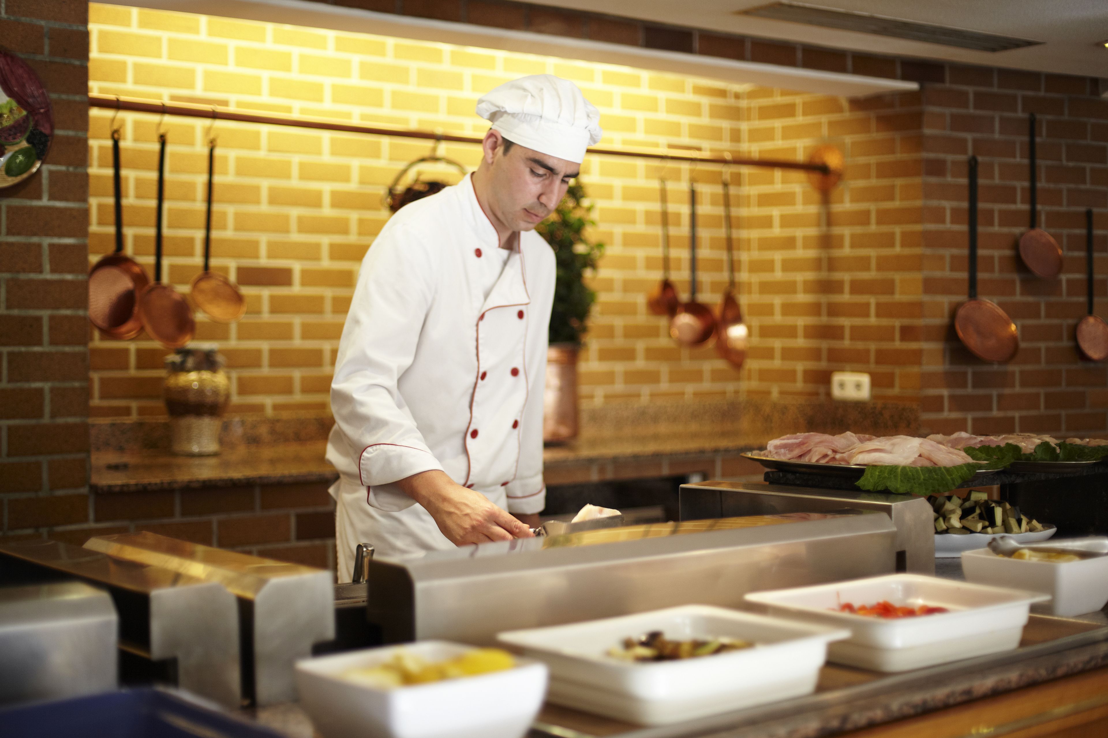 Un chef en una cocina con pared de ladrillo, ollas de cobre y varios recipientes de comida.