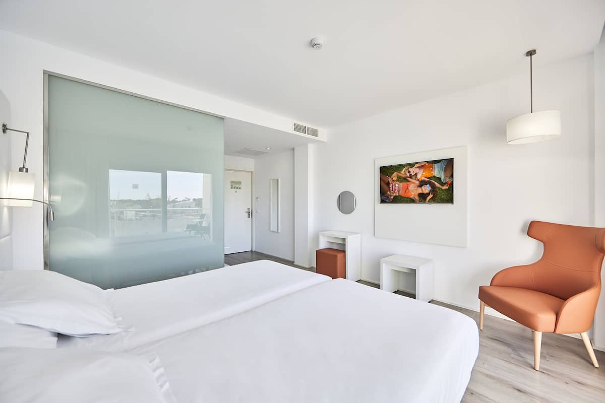 Une chambre d’hôtel moderne avec un grand lit, une cloison en verre, une petite table, une chaise et un tableau au mur.