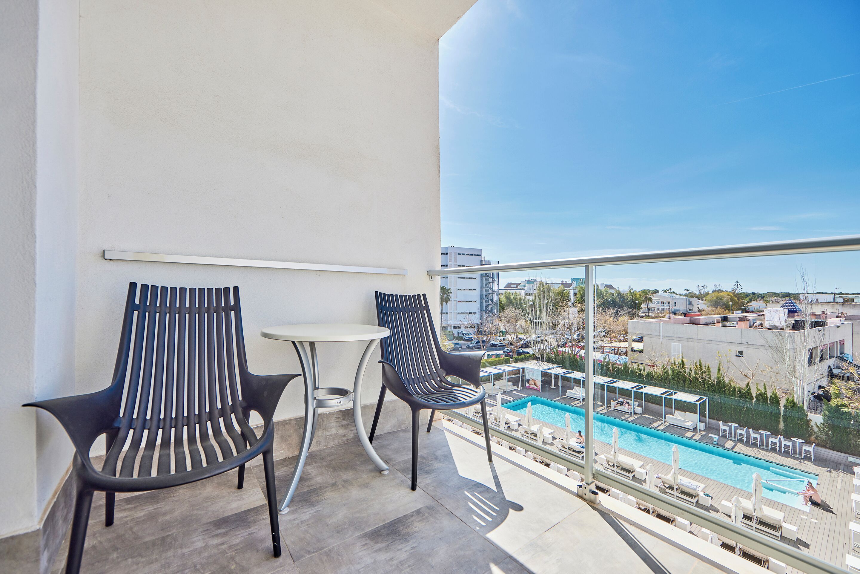 Un balcón con dos sillas y una mesita, con vista a un área de piscina con sillones de descanso y una vista despejada del paisaje urbano.
