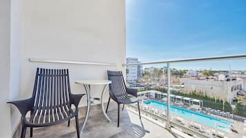 Un balcón con dos sillas y una mesita, con vista a un área de piscina con sillones de descanso y una vista despejada del paisaje urbano.