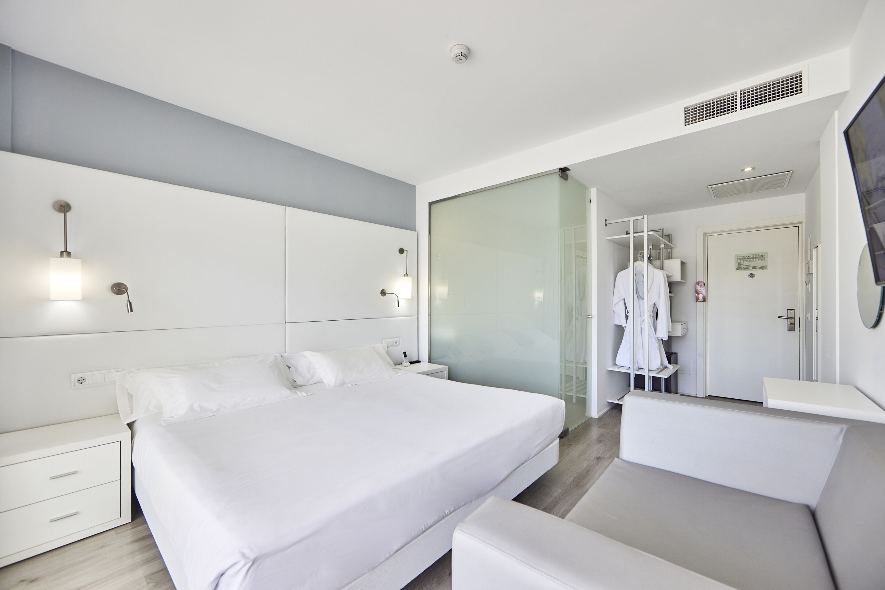 A modern hotel bedroom with a large bed, bedside tables, a wardrobe, and a glass partition.